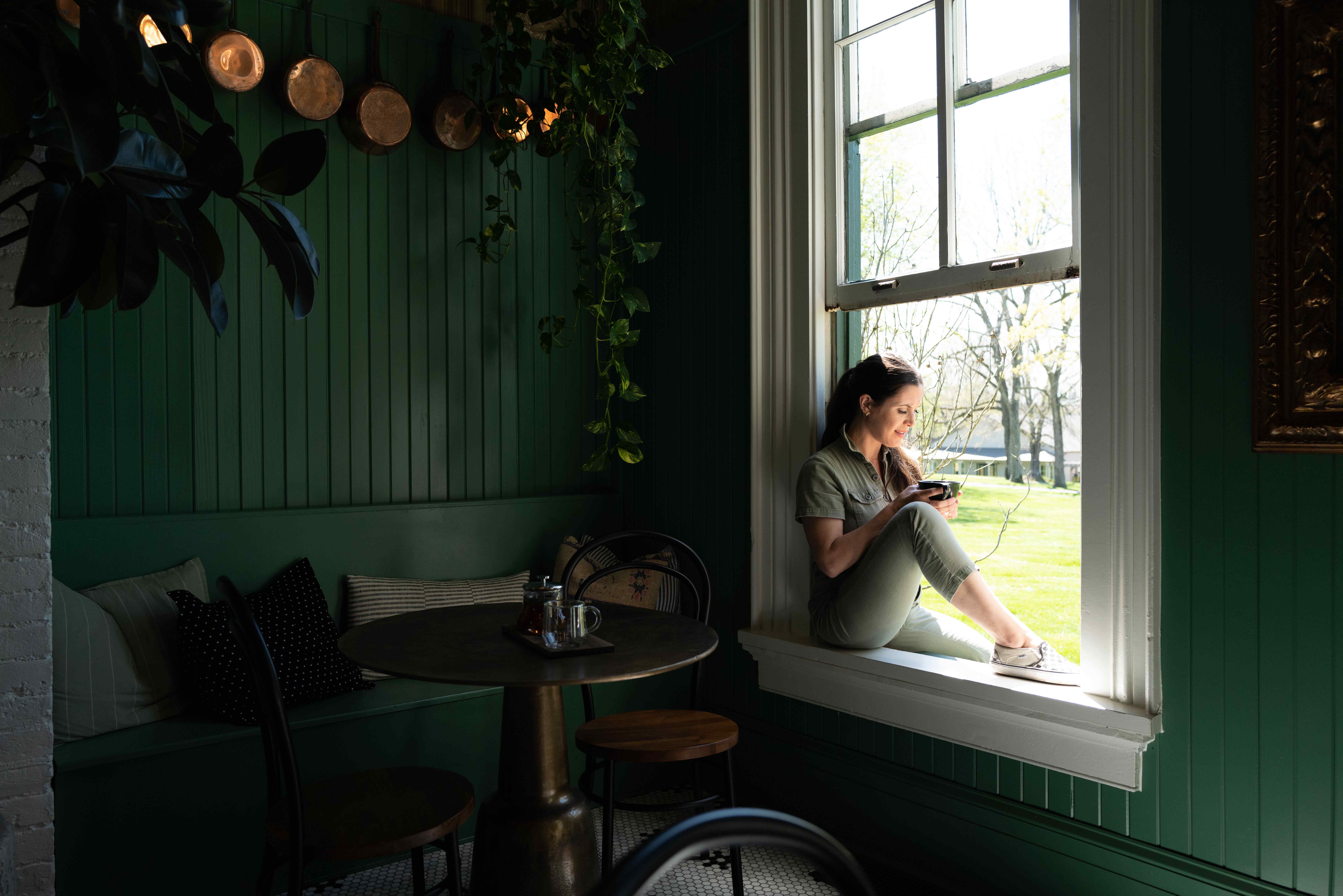 Woman sitting on a windowsill in a cozy green cafe, holding a smartphone, with natural light illuminating the space, reflecting a relaxed atmosphere for online wellness courses.