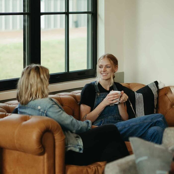 Two women engaging in a supportive conversation on a leather couch, one holding a cup, in a cozy, well-lit environment, reflecting the compassionate atmosphere of Milestones mental health treatment.